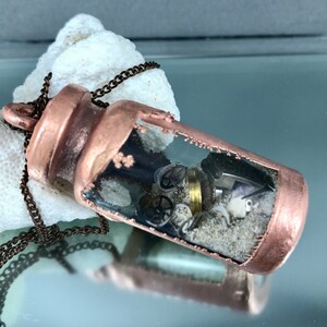 May include: A copper-colored pendant necklace with a glass vial containing sand, gears, and other small metal objects. The vial is attached to a brown chain.