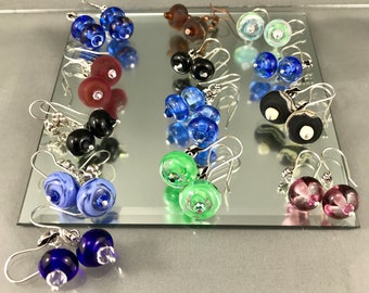 May include: Assortment of handmade glass earrings with silver hooks. The earrings feature various colors and patterns, including blue, green, purple, black, and white.