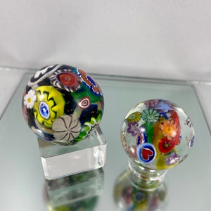 May include: Two colorful glass marbles with intricate patterns of flowers, hearts, and swirls. The marbles are displayed on small clear stands.