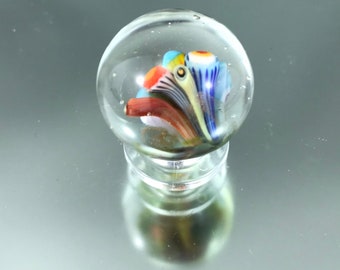 May include: A clear glass paperweight with a colorful, abstract glass design inside. The design features swirls of blue, red, yellow, and white glass.