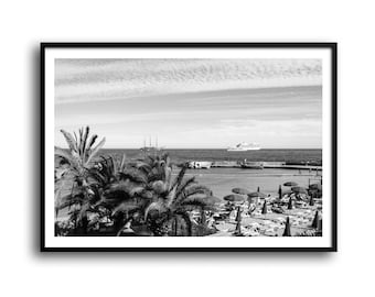 May include: Black and white framed print of a beach scene. Palm trees are in the foreground, with a beach, ocean, and boats in the background. The sky is filled with clouds. Beach umbrellas and lounge chairs are visible.