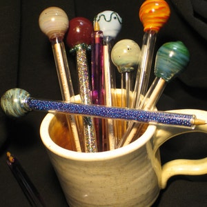 May include: A collection of colorful glass pens with different designs and patterns, including a blue and white swirl, a red and white swirl, and a purple pen with glitter, are displayed in a ceramic mug.