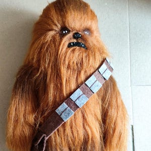 Könnte beinhalten: Ein Chewbacca-Plüschtier mit braunem und orangefarbenem Fell. Die Puppe hat ein detailliertes Gesicht mit schwarzen Augen und einer schwarzen Nase. Ein brauner Bandolier mit grauen Quadraten verläuft über die Brust. Die Puppe ist eine Star Wars-Figur.