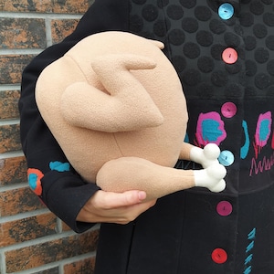 May include: A plush toy shaped like a roast chicken. The toy is beige with white feet and is being held by a person wearing a black coat with colourful embroidery.