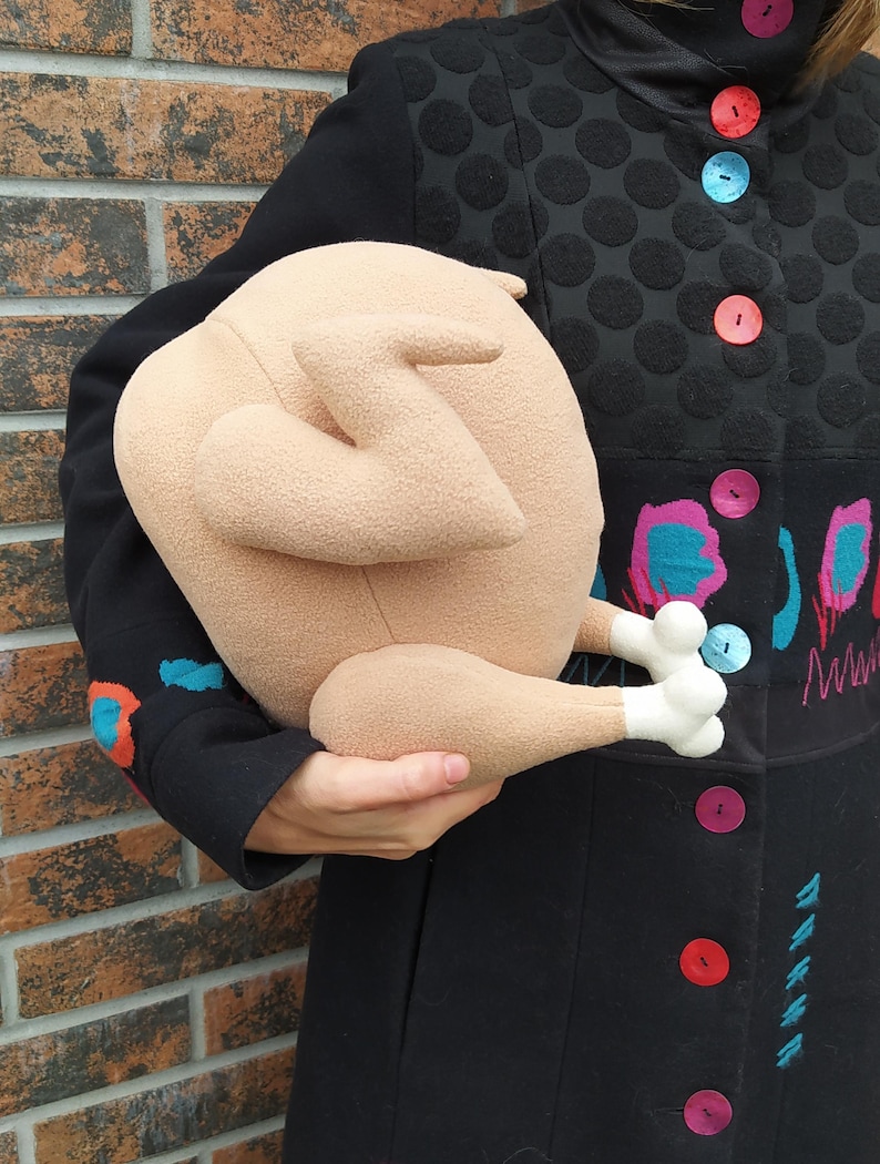May include: A plush toy shaped like a roast chicken. The toy is beige with white feet and is being held by a person wearing a black coat with colourful embroidery.