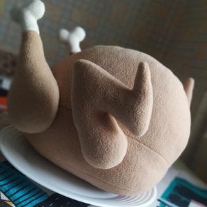 May include: A brown plush toy shaped like a roasted chicken. The chicken is lying on its side on a white plate. The toy has a realistic appearance with detailed feathers and a white bone sticking out of the leg.