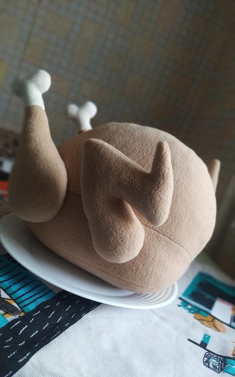 May include: A brown plush toy shaped like a roasted chicken. The chicken is lying on its side on a white plate. The toy has a realistic appearance with detailed feathers and a white bone sticking out of the leg.