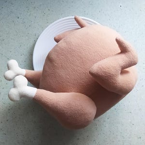 May include: A plush toy shaped like a roasted turkey, with a light brown colour and white feet. The turkey is lying on a white plate.