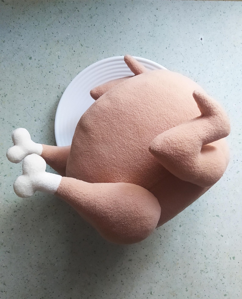 May include: A plush toy shaped like a roasted turkey, with a light brown colour and white feet. The turkey is lying on a white plate.