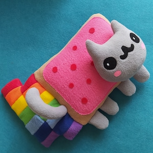 Puede incluir: Un gato de peluche gris con un cuerpo en forma de tostada rosa y marrón. El gato tiene una cola de arcoíris y una expresión feliz.