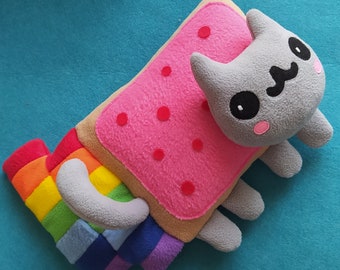 Puede incluir: Un gato de peluche gris con un cuerpo en forma de tostada rosa y marrón. El gato tiene una cola de arcoíris y una expresión feliz.