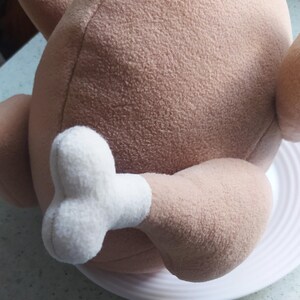 May include: A plush toy shaped like a roasted turkey with a white leg and foot. The turkey is brown and has a soft, fuzzy texture.