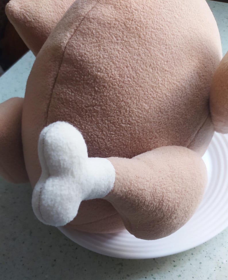 May include: A plush toy shaped like a roasted turkey with a white leg and foot. The turkey is brown and has a soft, fuzzy texture.