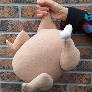 May include: A plush toy shaped like a roast chicken. The chicken is brown and has white feathers on the ends of its wings and legs.