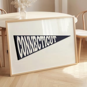May include: Framed wall art showing a navy blue pennant with the word "CONNECTICUT" in white, bold, retro lettering. The artwork is in a light wood frame and leans against a wall. The background includes a wooden table and chairs.