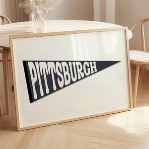 Puede incluir: Impresión enmarcada con un banderín azul marino con la palabra "PITTSBURGH" en letras blancas. La impresión está en un marco de madera clara. El fondo incluye una mesa y sillas de madera.
