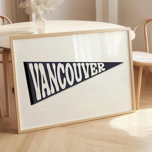 May include: A framed print featuring a navy blue pennant with the word "VANCOUVER" in bold, white, retro-style lettering. The print is displayed in a light wood frame, leaning against a wall. The overall aesthetic is minimalist and modern.