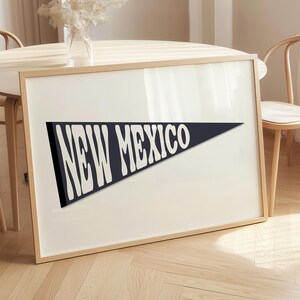 May include: A framed print featuring a navy blue pennant with the words "NEW MEXICO" in a white, stylized font. The print is set against a cream-colored background within a light wood frame. The artwork is displayed on a wooden floor, near a round table and chairs.