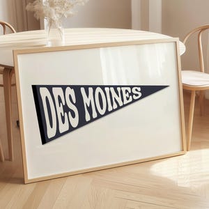 Könnte beinhalten: Gerahmter Druck im Wimpel-Stil mit dem Text "DES MOINES" in Weiß auf marineblauem Hintergrund. Der Rahmen ist aus hellem Holz. Der Druck liegt auf einem Tisch, mit Stühlen im Hintergrund, in einem hellen Raum.