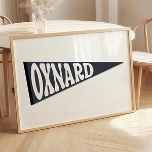 May include: A framed print featuring the word "OXNARD" in white, stylized lettering on a navy blue pennant shape. The print is set against a cream-colored background within a light wood frame. The artwork is leaning against a table.
