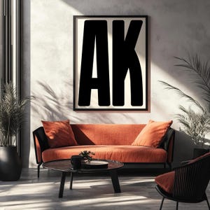 Puede incluir: Un salón moderno con un sofá naranja quemado y cojines a juego. Una obra de arte enmarcada en negro con las letras "AK" en negrita negra cuelga sobre el sofá. Una mesa de centro redonda y una silla negra completan la escena.