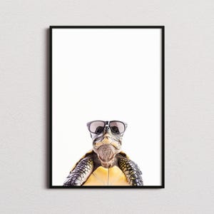 Op de afbeelding: Een ingelijste wandkunstprint met een schildpad die een zwarte zonnebril draagt. De schildpad heeft een getextureerd, patroonrijk schild en huid in tinten bruin, beige en geel. Het onderwerp is gecentreerd tegen een effen witte achtergrond.