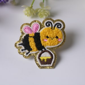May include: A gold glitter iron-on patch featuring a cartoon bee carrying a honey pot. The bee is yellow with black stripes and pink wings.