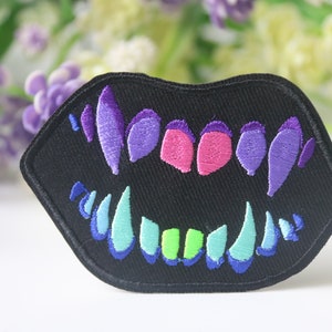 May include: Black embroidered patch featuring a stylized mouth with colorful teeth. The teeth are in shades of purple, pink, green, and blue. The patch has a black border and is likely designed to be ironed or sewn onto fabric.