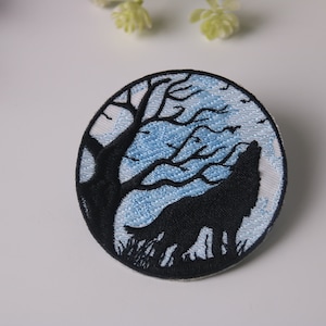 Peut inclure: Patch brodée thermocollante représentant un loup noir hurlant à la lune, en silhouette sur un fond de lune bleu et blanc et un arbre dénudé.