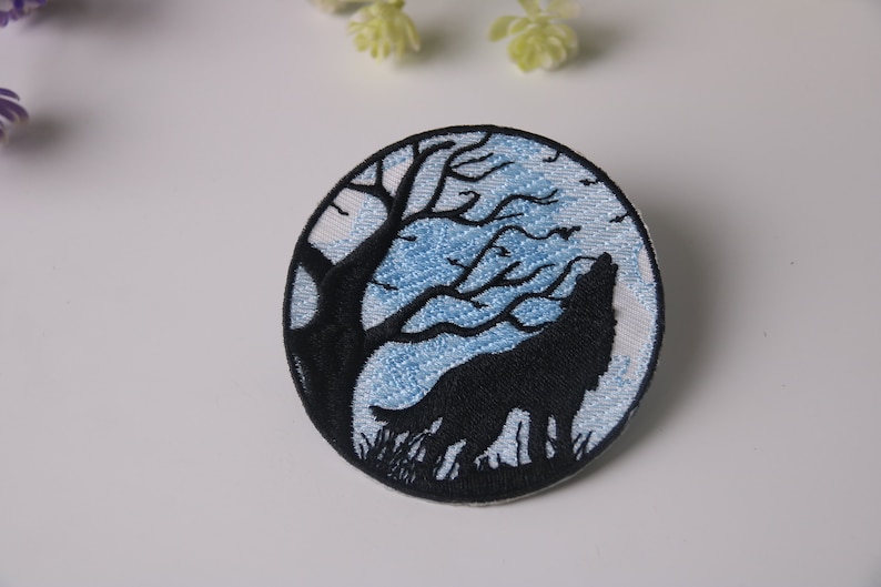 Op de afbeelding: Geborduurde strijkpatch met een zwarte wolf die huilt naar de maan, gesilhouetteerd tegen een blauwe en witte maan en een kale boom.