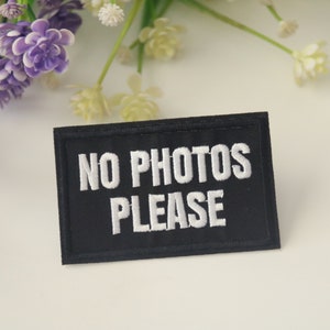 May include: Black fabric patch with white embroidered text that reads "NO PHOTOS PLEASE".