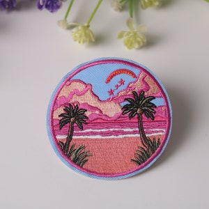 May include: A round embroidered patch depicting a pink and blue sunset scene. Two palm trees stand on a pink beach with a blue ocean. The sky is blue with pink clouds and a red arc. There are three white stars in the sky.