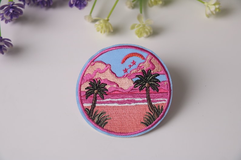 Peut inclure: Un patch rond brod&eacute; repr&eacute;sentant une sc&egrave;ne de coucher de soleil rose et bleu. Deux palmiers se dressent sur une plage rose avec une mer bleue. Le ciel est bleu avec des nuages roses et un arc rouge. Il y a trois &eacute;toiles blanches dans le ciel.