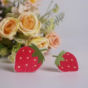 Könnte beinhalten: Zwei gestickte Erdbeer-Patches mit roten Körpern und grünen Stielen. Die Patches haben weiße Ränder und kleine weiße Samendetails. Im Hintergrund ein Strauß aus pfirsichfarbenen und rosa Blumen.