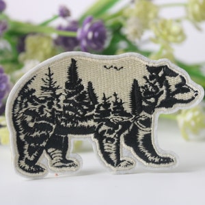 Op de afbeelding: Geborduurde patch in de vorm van een beer, met een zwart boslandschap tegen een beige achtergrond. De beer is wit omlijnd, met een klein vogeldetail boven de bomen. Ideaal voor jassen, tassen of andere stoffen items.