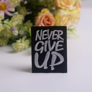 K&ouml;nnte beinhalten: Ein schwarzer rechteckiger Aufn&auml;her mit den Worten "NEVER GIVE UP" in Wei&szlig; gestickt. Der Aufn&auml;her befindet sich vor einem verschwommenen Hintergrund aus Blumen in Rosa-, Gelb- und Gr&uuml;nt&ouml;nen.
