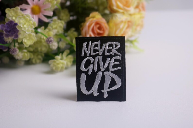 K&ouml;nnte beinhalten: Ein schwarzer rechteckiger Aufn&auml;her mit den Worten "NEVER GIVE UP" in Wei&szlig; gestickt. Der Aufn&auml;her befindet sich vor einem verschwommenen Hintergrund aus Blumen in Rosa-, Gelb- und Gr&uuml;nt&ouml;nen.