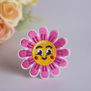 May include: Pink flower embroidered patch with a yellow smiley face in the center. The smiley face has black eyes, a small mouth, and red cheeks.