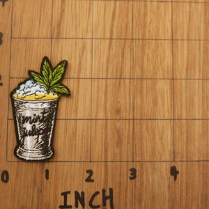 Op de afbeelding: Geborduurde patch met een cocktailglas met de tekst "mint julep". Het glas is gevuld met een wit en blauw mengsel, bekroond met een groen blad. De patch is ongeveer 5 cm hoog.
