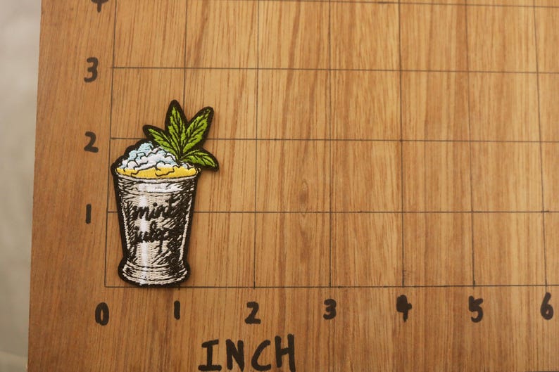 Op de afbeelding: Geborduurde patch met een cocktailglas met de tekst "mint julep". Het glas is gevuld met een wit en blauw mengsel, bekroond met een groen blad. De patch is ongeveer 5 cm hoog.