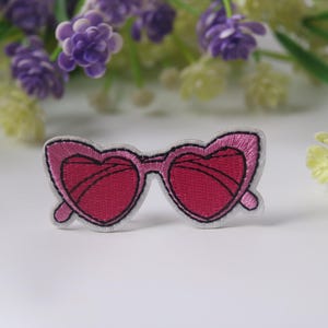 Puede incluir: Parche bordado de gafas de sol en forma de corazón rosa. El parche está sobre un fondo blanco.