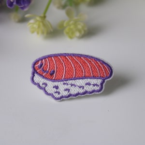 May include: A purple and white embroidered patch of a sushi roll with a pink salmon filling. The sushi has a cute cartoon face.