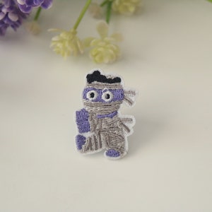 May include: An embroidered iron-on patch of a mummy wearing a purple mask and holding a purple bandage. The mummy is gray and white.