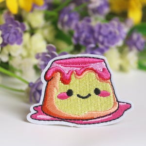 May include: A pink and yellow embroidered iron-on patch of a smiling pudding with a pink swirl on top.