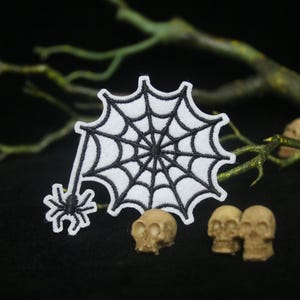 以下が含まれることがあります： 白い布地に白いクモの巣と黒いクモが刺繍されたパッチ。パッチは黒い背景に白い小さな頭蓋骨で囲まれています。