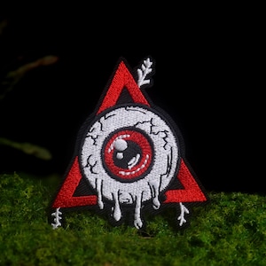 May include: An embroidered patch featuring a white, bloodshot eyeball with a red iris and black pupil, centered within a red triangle. The eyeball appears to be melting with white drips, and a small white arrow points upward from the top.