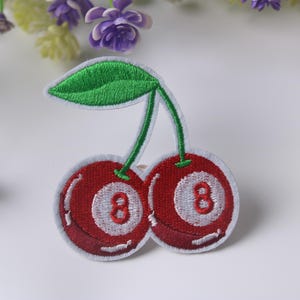 Könnte beinhalten: Gestickter Aufnäher mit zwei roten Kirschen, auf denen jeweils die weiße Zahl 8 zu sehen ist. Die Kirschen haben grüne Stiele und ein grünes Blatt. Für Kleidung oder Accessoires.
