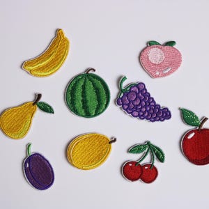 May include: A collection of embroidered fruit patches. The image shows a yellow banana, green watermelon, pink peach, purple grapes, yellow pear, red apple, red cherries, yellow lemon, and purple plum. These patches are suitable for decorating clothes or accessories.