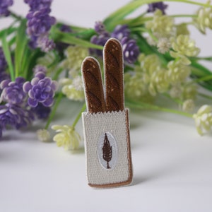 May include: A white embroidered patch featuring two brown baguette loaves in a paper bag. The bag has a small green leaf design on the front.