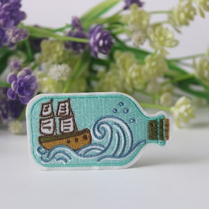 Può includere: Una toppa ricamata blu di una nave in una bottiglia con onde e bolle. La bottiglia ha un tappo di sughero.